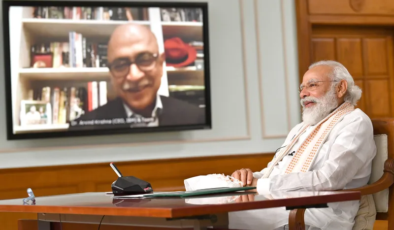 Prime Minister Narendra Modi seated at a desk, smiling at a large screen showing Arvind Krishna on a video call
