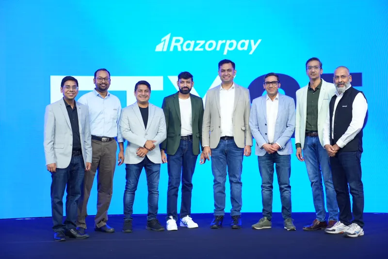 Eight Razorpay leaders standing on a stage in front of a blue screen with the Razorpay logo; Shashank Kumar in a dark green blazer and Harshil Mathur in a beige blazer stand fourth and fifth from left