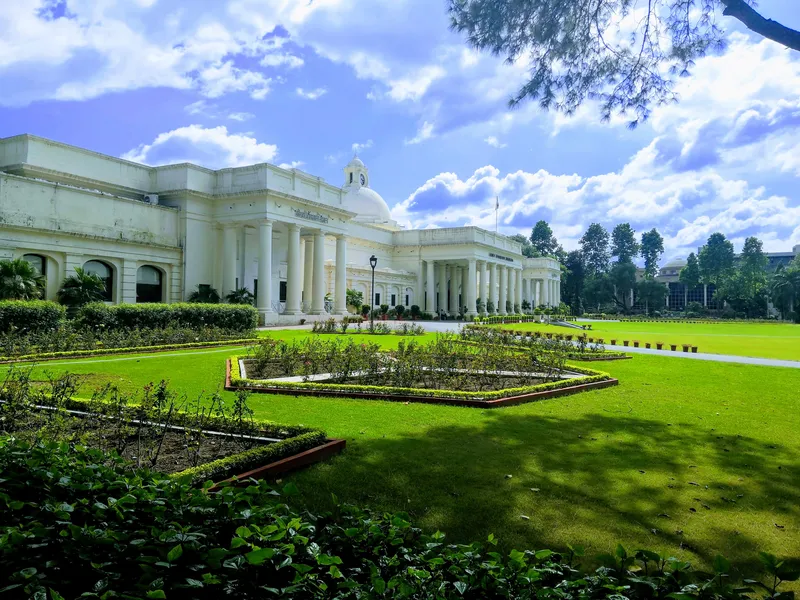 The white colonnaded main building of IIT Roorkee seen across green lawns and hedged flower beds under a blue sky