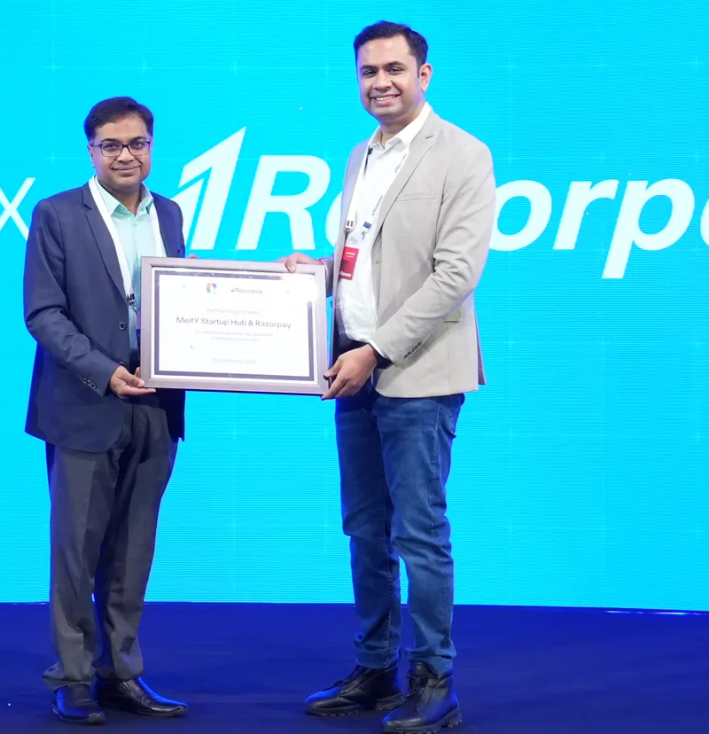 Harshil Mathur in a light grey blazer, white T-shirt and jeans holds a framed certificate with Panneerselvam Madanagopal of MeitY Startup Hub, in a navy suit, on a stage with a cyan screen