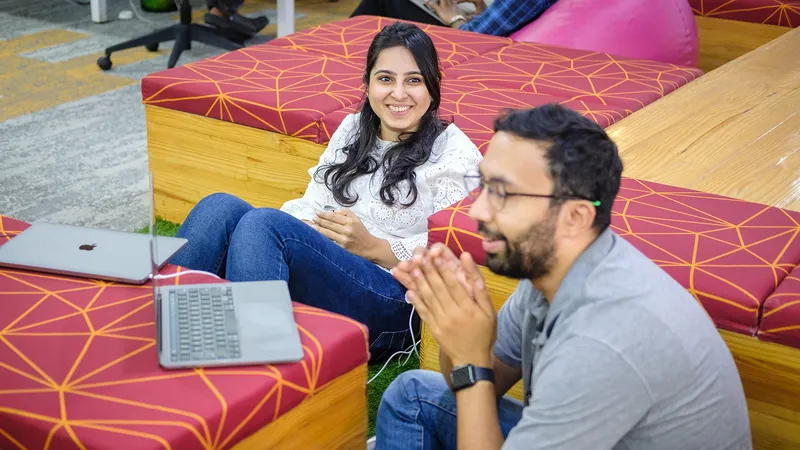 Two Razorpay employees with laptops sitting on red geometric-patterned seating in an office lounge