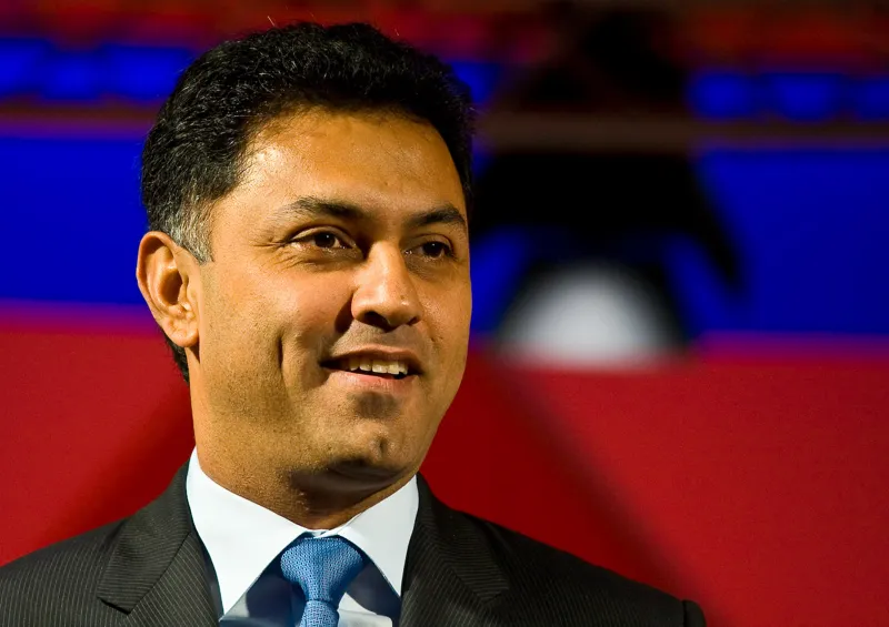 Nikesh Arora in a dark suit and blue tie, smiling, against a red and blue stage backdrop